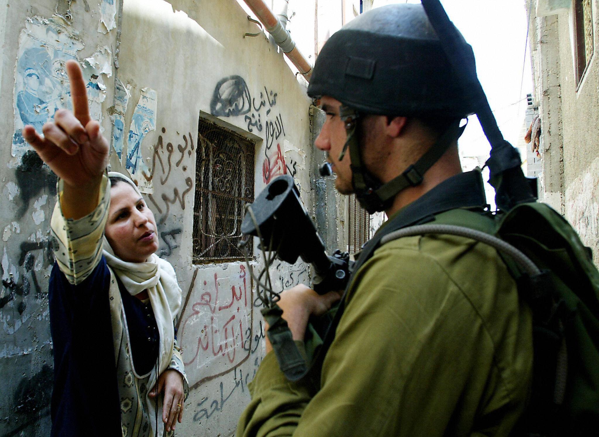 A Palestinian woman raises her hand while confronting an armed Israeli soldier in an alleyway marked with political graffiti.